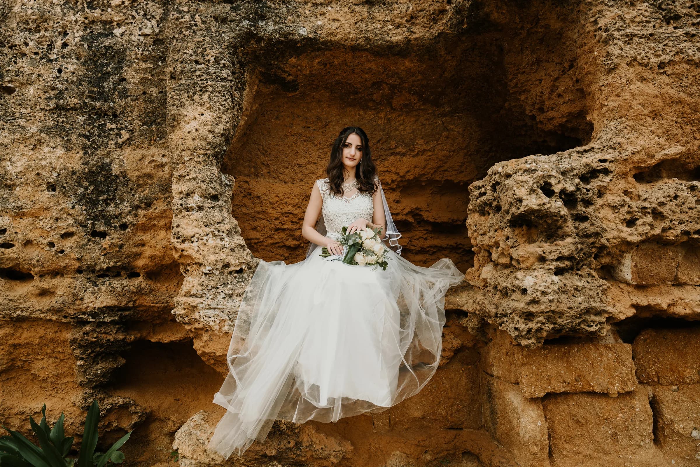 A bride seated in a golden stone alcove, her dress across the rock